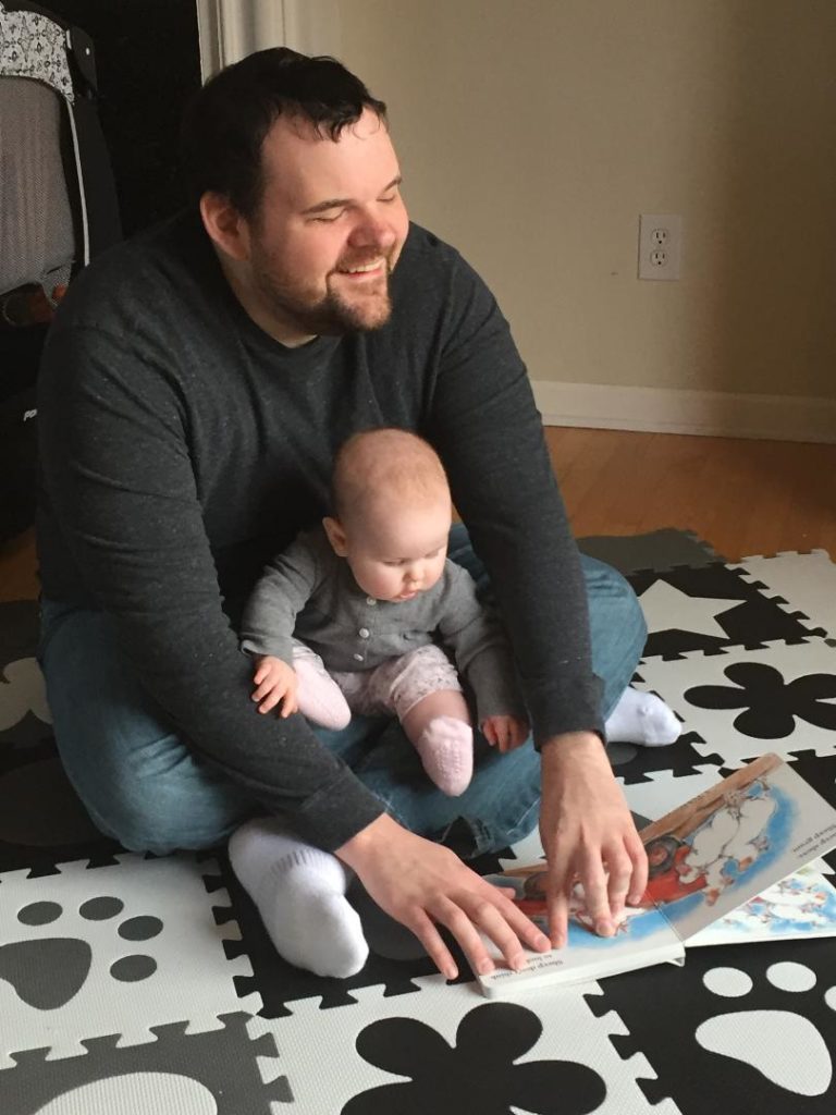 Jared and his baby daughter Harper sit on the floor while he reads a braille book to her.