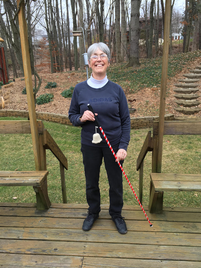 Carol stands on a wooden deck outdoors smiling broadly and holding a red-and-white-striped mobility cane that has a bell affixed to the handle.