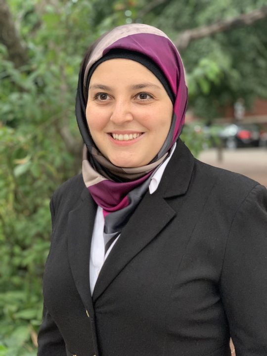 Mona, standing outdoors, wearing a colorful hijab and a black suit jacket, smiles for the camera.