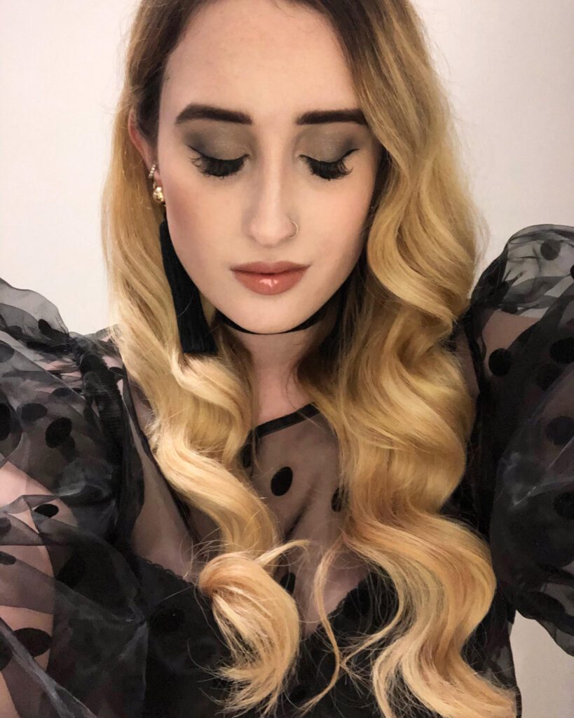 A selfie of blind painter, Kimberley Burrows. She has long, wavy, golden blonde hair. and is wearing black tasselled earrings, a black choker, and a black sheer shirt with ruffled shoulders that is covered in large polka dots.
