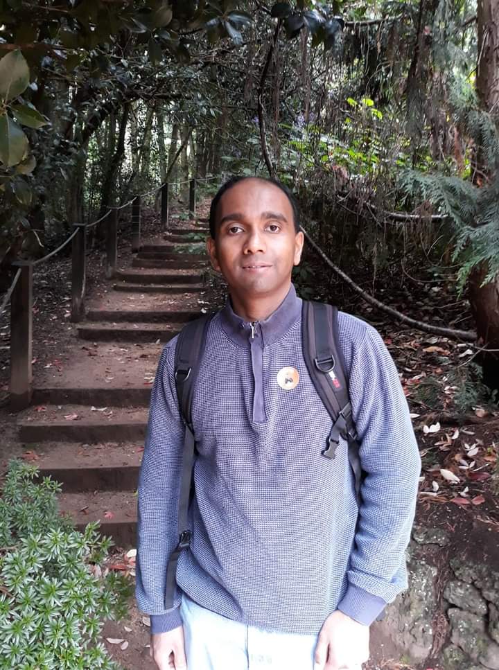 Prabath, wearing hiking gear, stands in a tropical forest at the base of a set of steps that lead up a trail.
