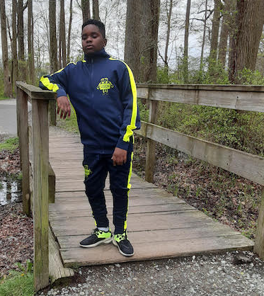 Isaiah stands on a wooden bridge in a forest wearing one of his company’s tracksuits and looking confidently into the camera.