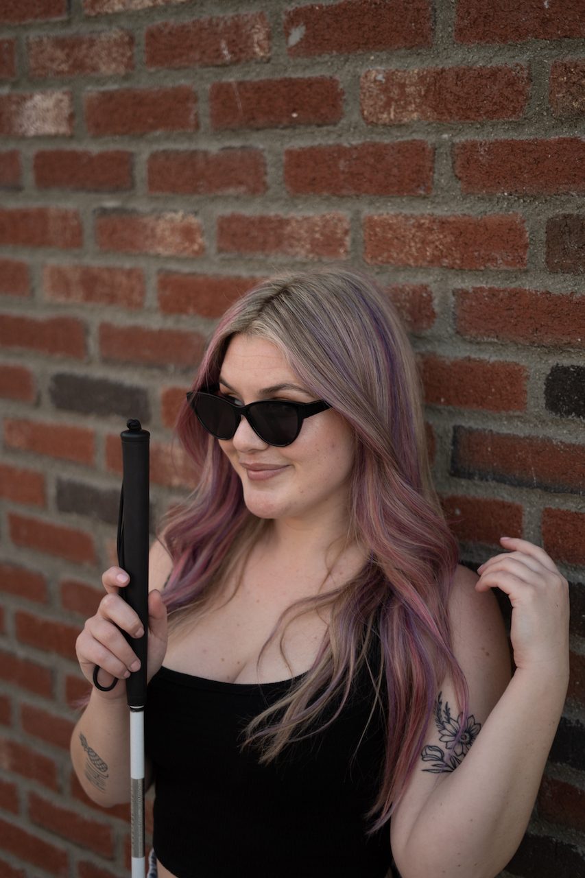 Jessie stands against a brick wall wearing dark glasses and holding her mobility cane, smiling