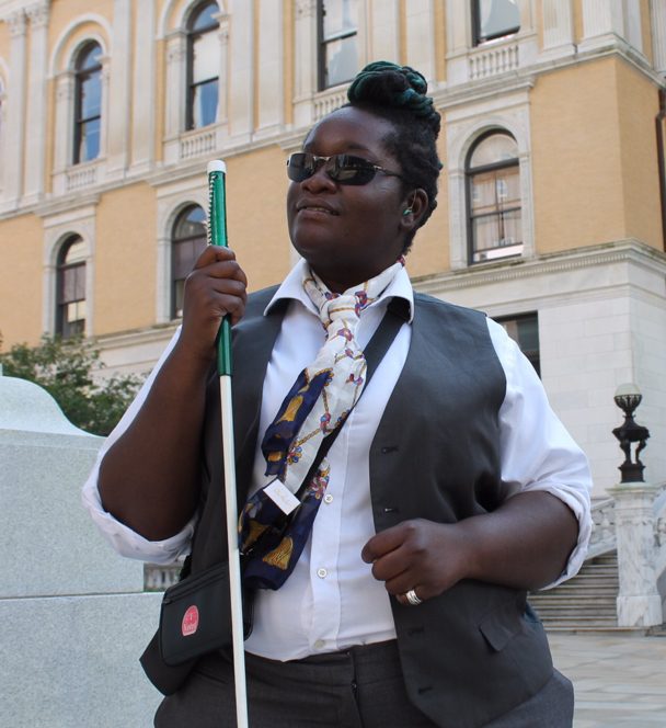 A Black woman stands confidently on a city street, holding her mobility cane in her fist.