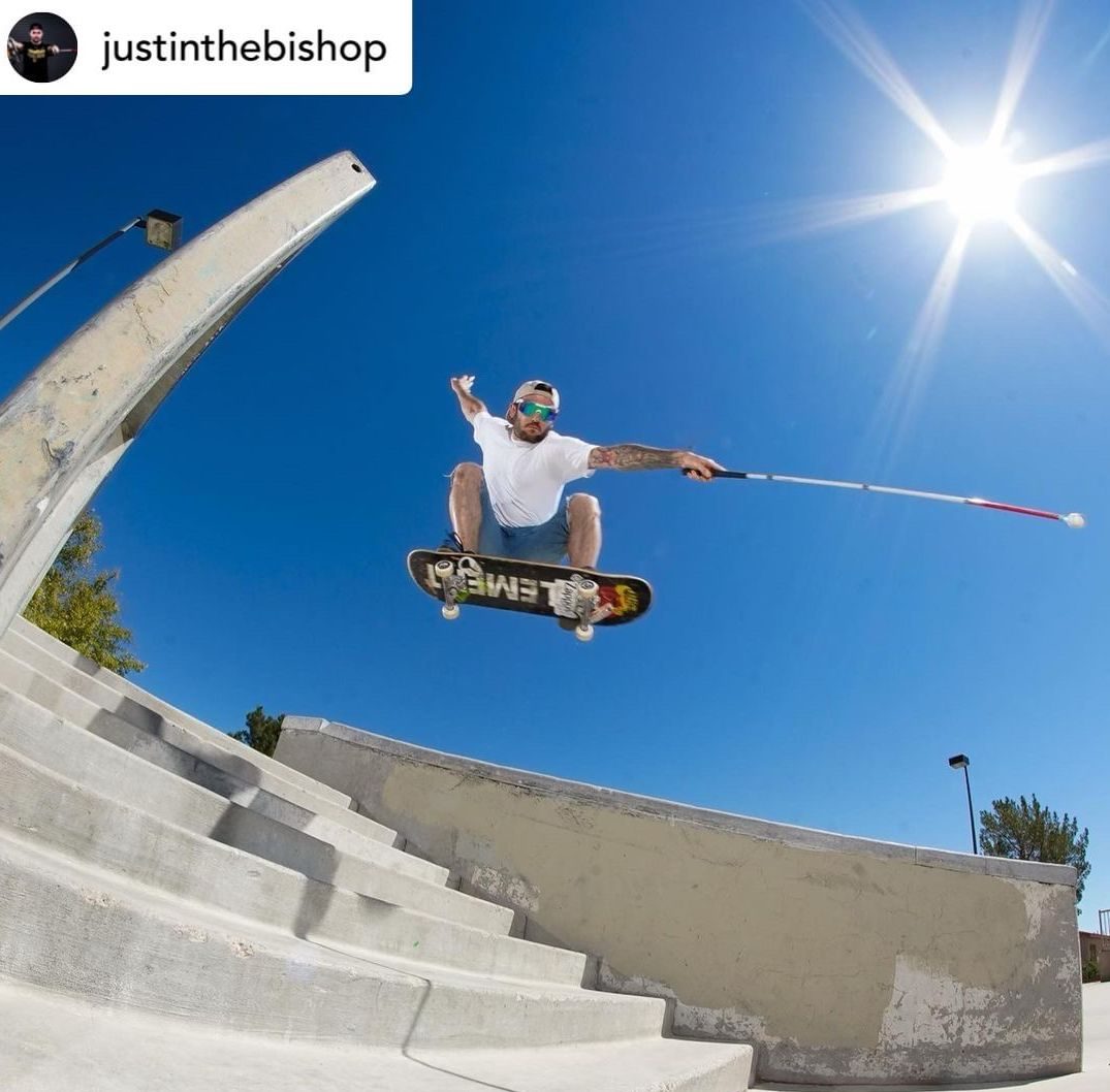 Against a bright blue sky and blazing sun, pro skateboarder Justin Bishop holds his mobility cane out in the air as he flies over a set of seven stairs, doing an "ollie."