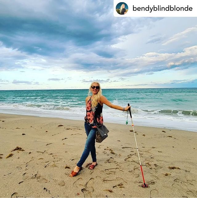 Monica at the beach, wearing dark glasses, smiles and leans into her mobility cane on the sand while waves crash behind her and gray clouds gather overhead.