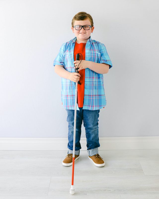 Karl is smiling and posed in front of a white wall while holding a white cane.