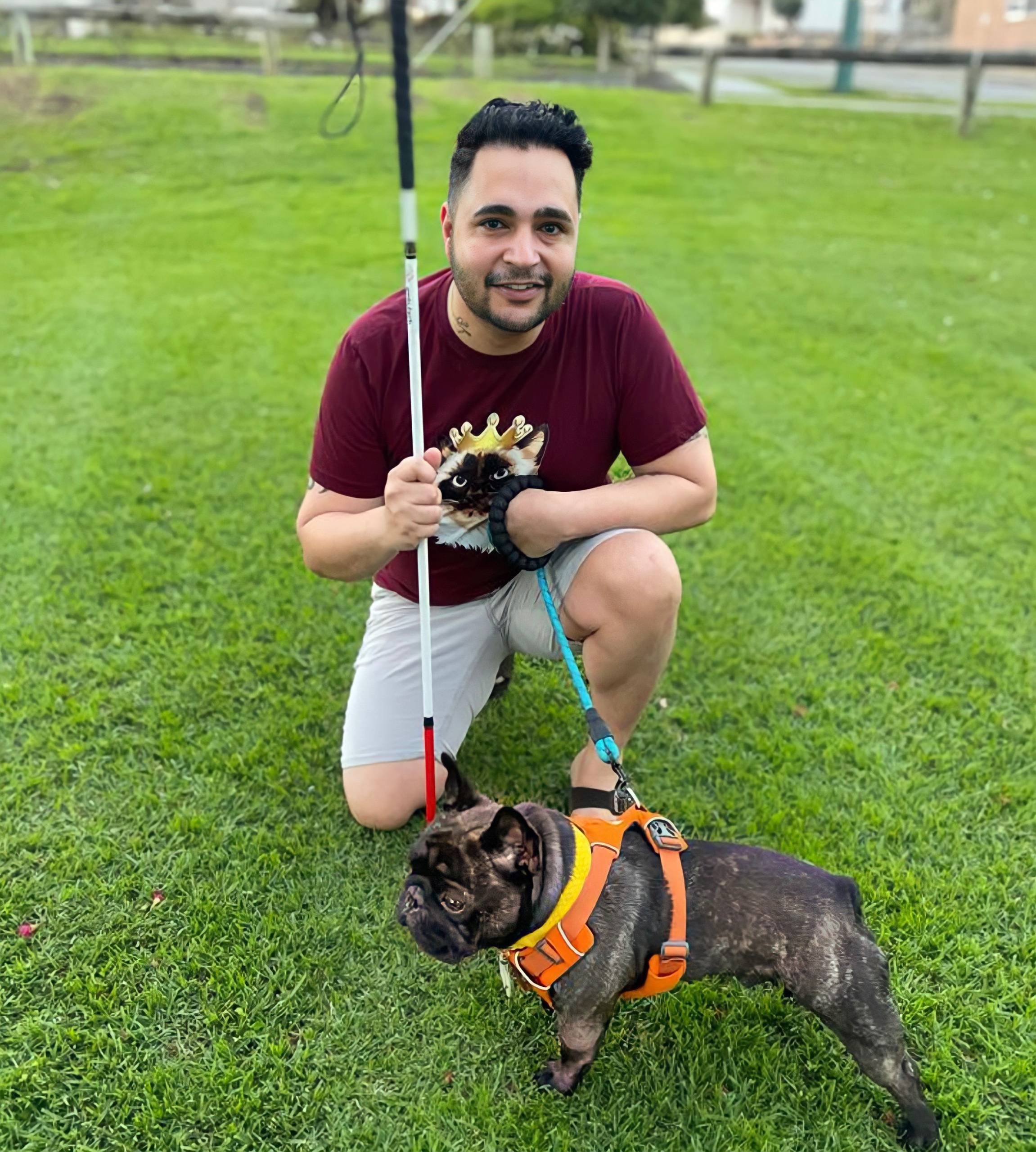 Karan Nagrani kneels on the grass holding his mobility cane in one hand and the leash for Henry, his small French bulldog, in the other.