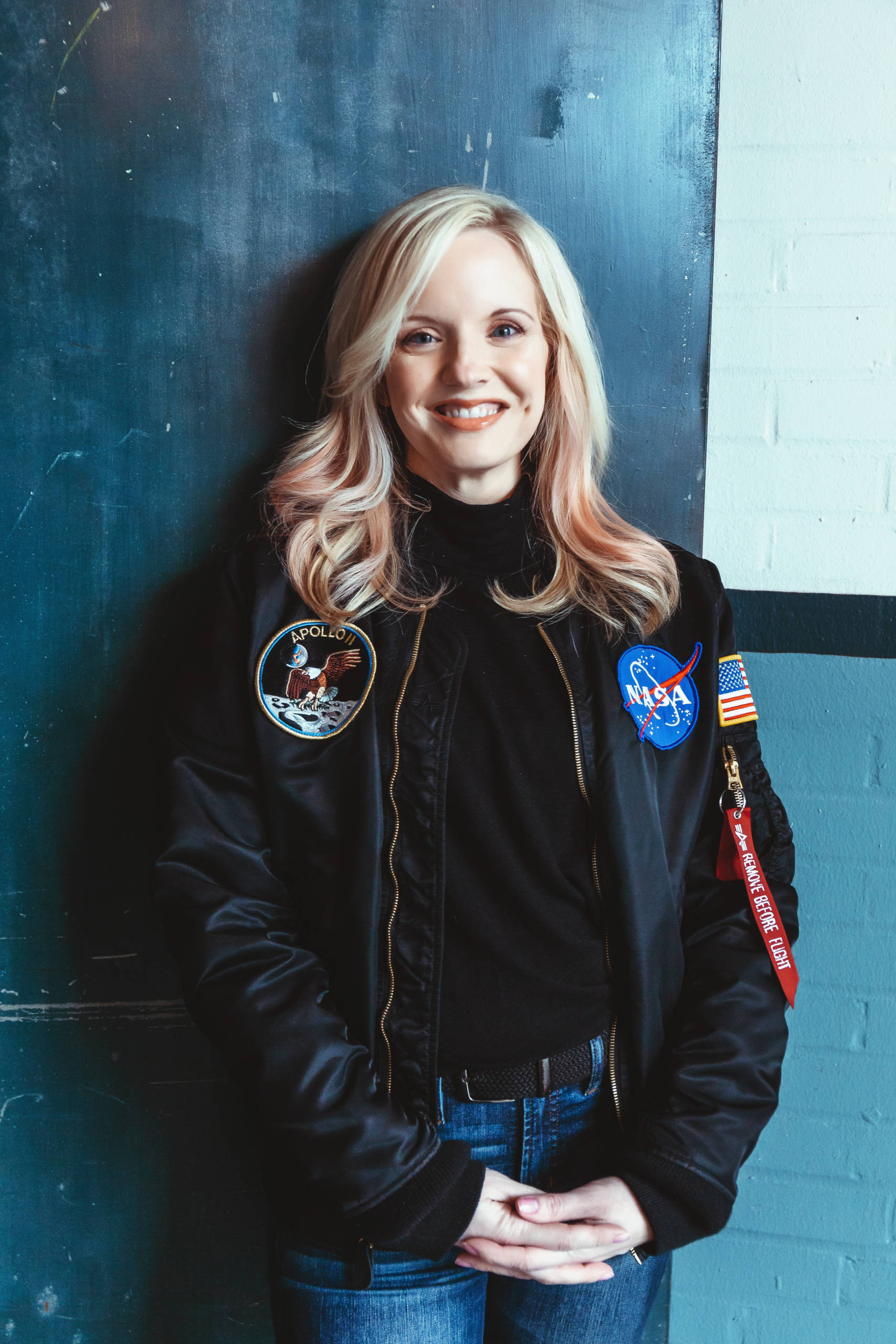 Kim Arcand stands against a wall smiling, wearing a NASA jacket