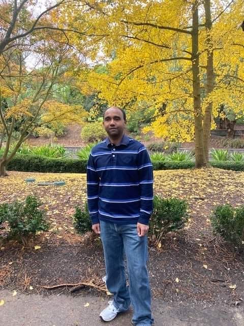Prabath standing on a path in a park, surrounded by trees with yellow leaves, many of which have fallen to the ground.