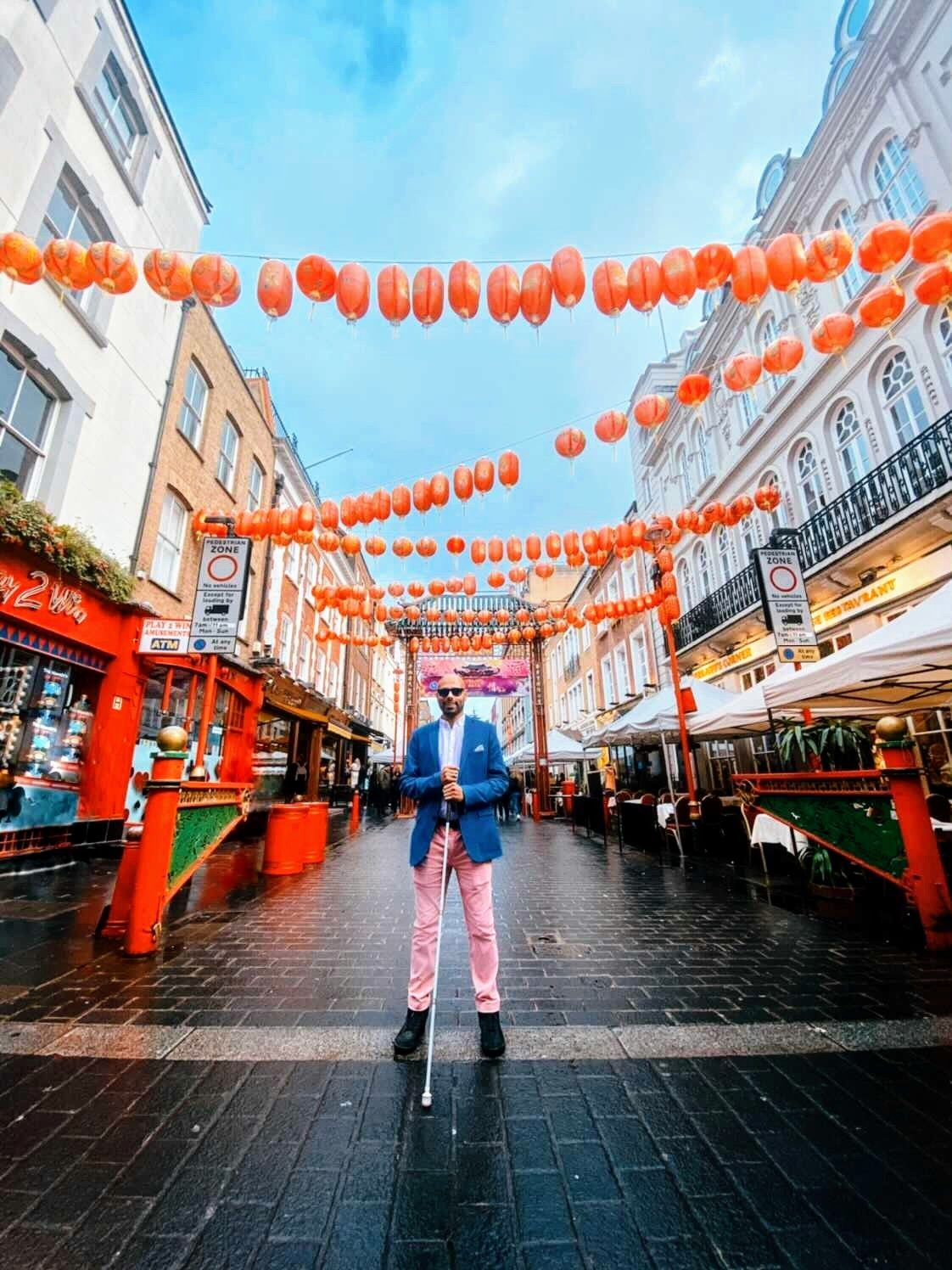 Yahya, a sharply dressed young man wearing pink pants, a blue blazer and dark glasses, holding his mobility cane in the middle of a cobblestone street decorated with bright paper lanterns.