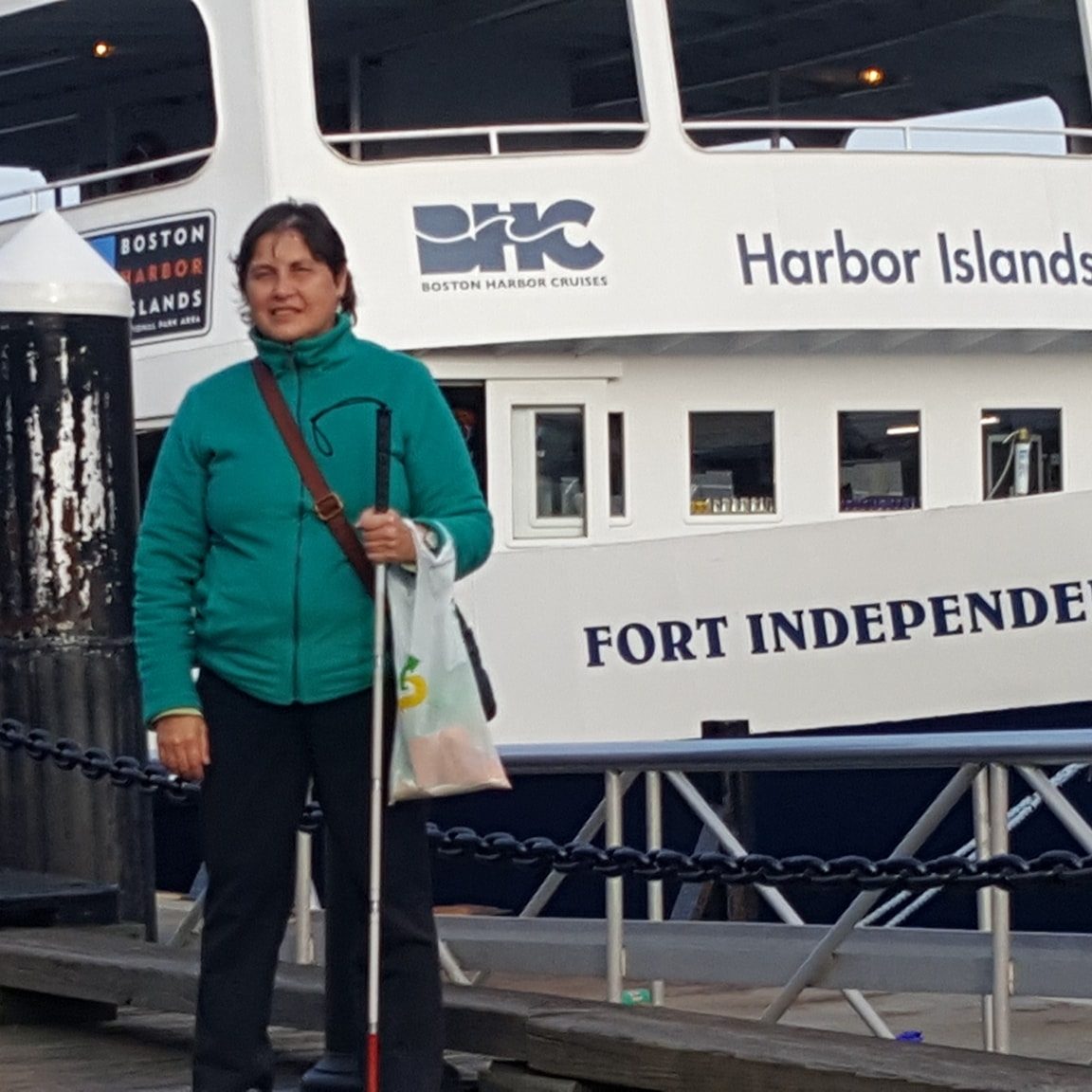 Christine, smiling and holding her mobility cane, stands in front of the Fort Independence boat at Boston Harbor Cruises.