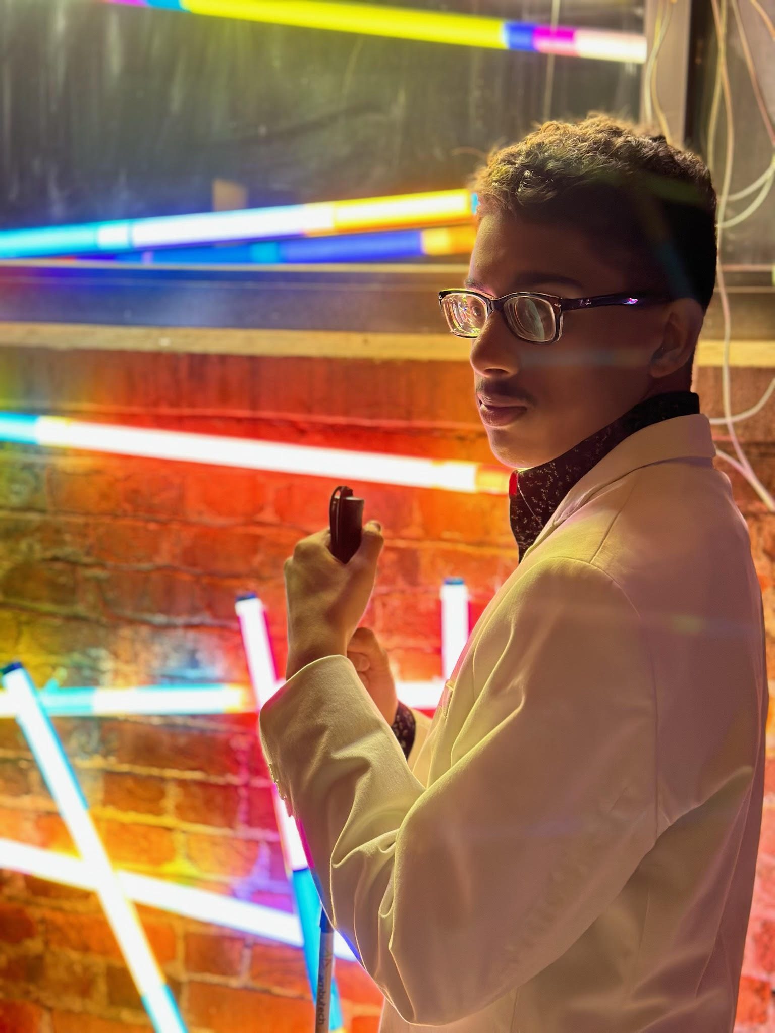 Caiden, a young man wearing glasses and a white suit, stands in front of an art installation of multi-colored neon lights while holding his mobility cane.