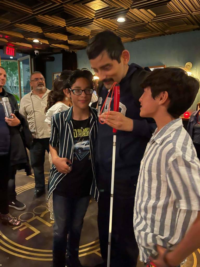 Blind actor Matthew Saracho stands in the lobby of a theater, holding his mobility cane and chatting with two young people.