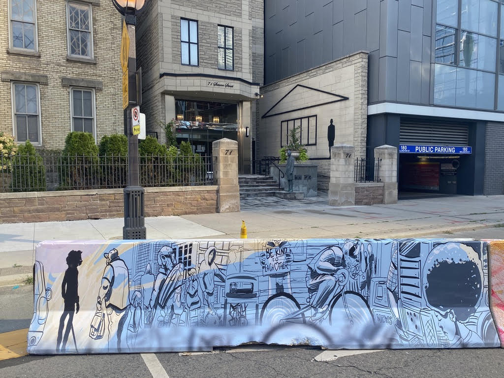 A painting on a cement traffic barrier: a silhouette of a blind gentleman in a very busy city setting, a man walking his child, an old couple walking, a cyclist zooming by and a girl checking her phone. (Photo credit: Phil Sutherland)