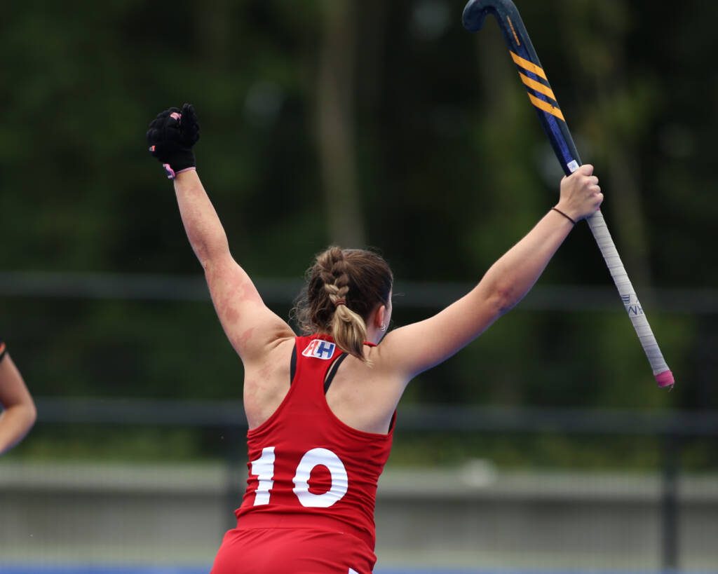 A player wearing a number 10 jersey faces away from the camera, holding her field hockey stick high in the air in victory