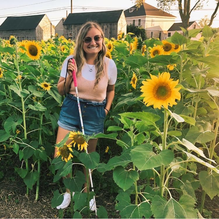 Frankie Ann stands among towering sunflowers, holding her mobility cane and smiling broadly for the camera