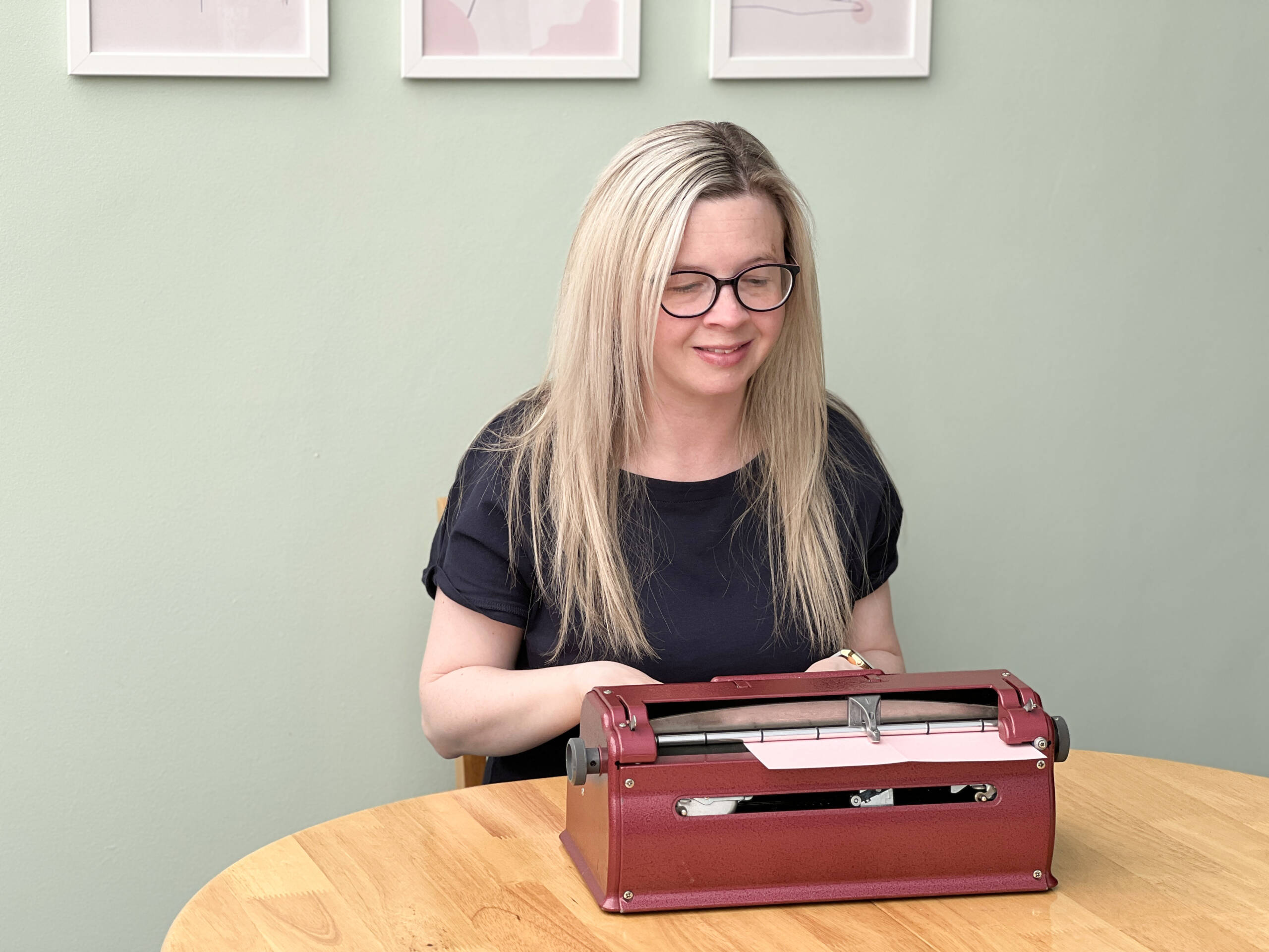 Owner of Dotty About Braille Hayley Kellard, a white woman with long, blonde hair wearing glasses, sits at a red Perkins Brailler working on a card