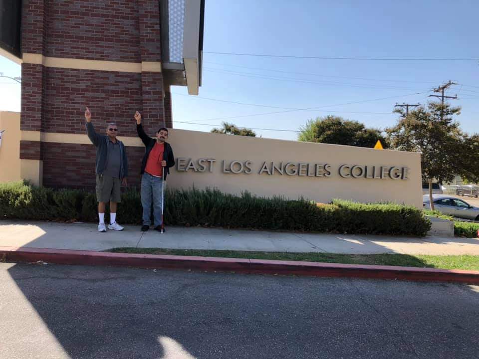 Matthew Saracho, holding his white mobility cane, and a companion in front of a sign for East Los Angeles College with their arms up in victory
