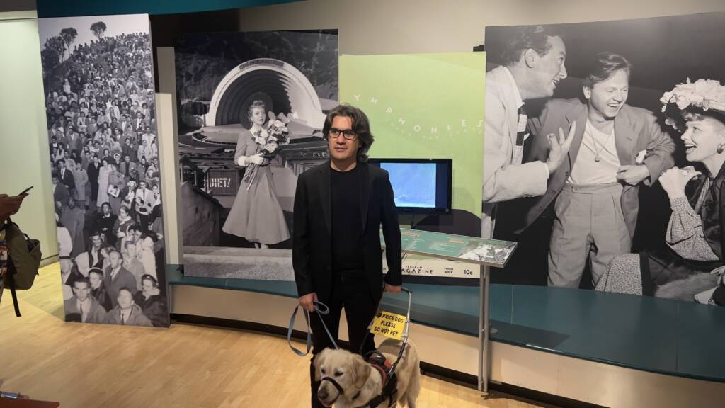 Composer Kemal Gorey and his guide dog stand in front of an exhibit labeled "Symphonies Under the Stars," featuring black-and-white, mid 20th-century photos of people at the Hollywood Bowl.