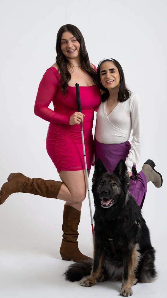 Juna Gjata and Lina Coral, hosts of the Blind Girl Chat podcast. Juna is wearing a hot pink dress and holding a white cane. Lina is wearing a white shirt and long purple skirt and holding the least for her guide dog, a German Shepherd named Quest.