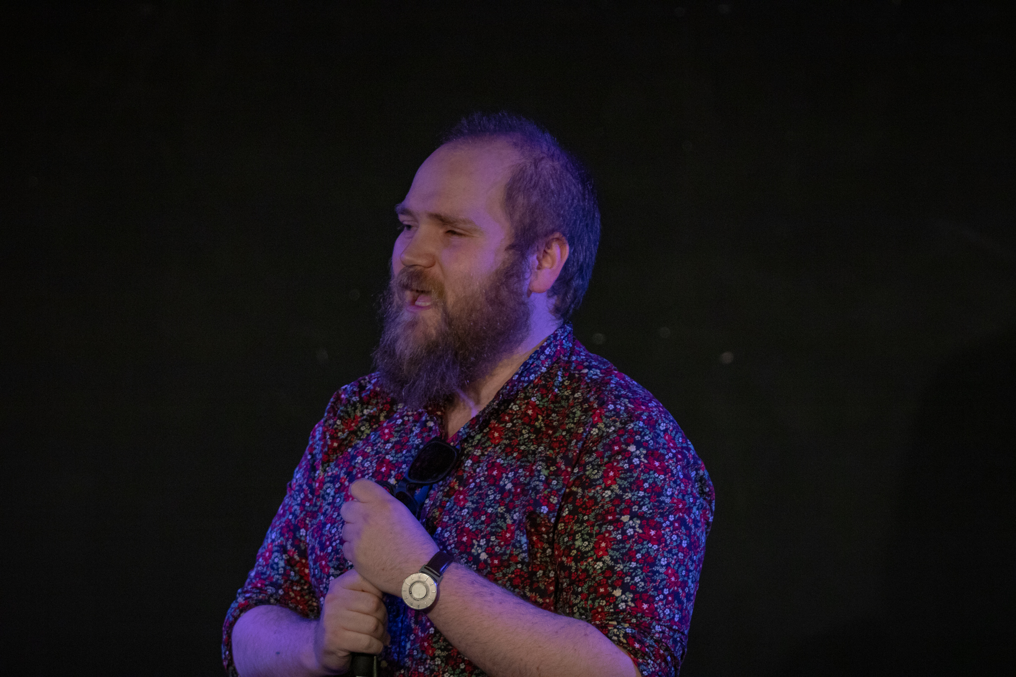 Extant Theatre's Ben Wilson, a white man with short blonde hair and a beard wearing a floral shirt, is captured on-stage, mid-sentence. (Credit: Jamie Dennis, Primo Digital Video Productions)