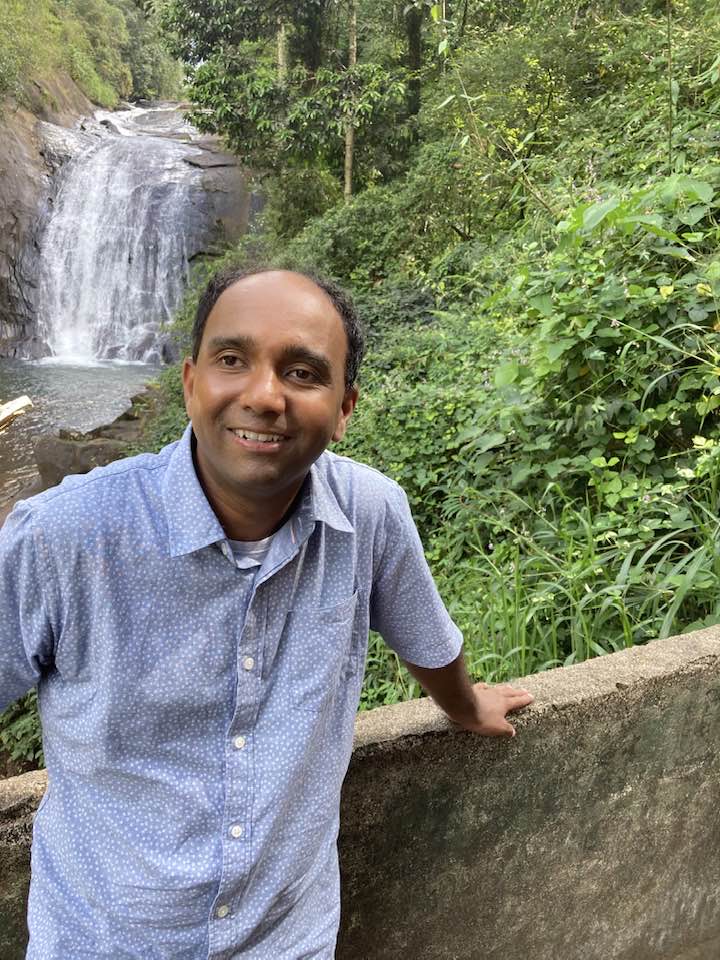 Inclusion advocate Prabath Wickramanayake stands smiling among lush greenery in front of a small waterfall