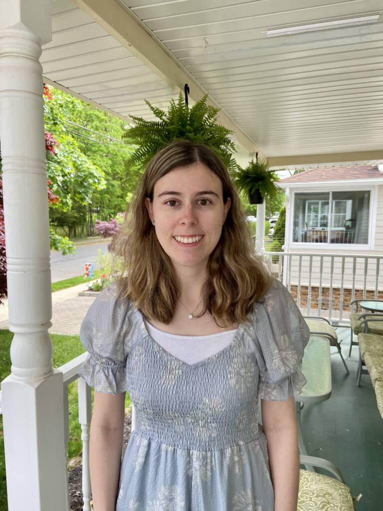 Marianne, a white woman with long, light brown hair, stands on a sunny porch, smiling for the camera