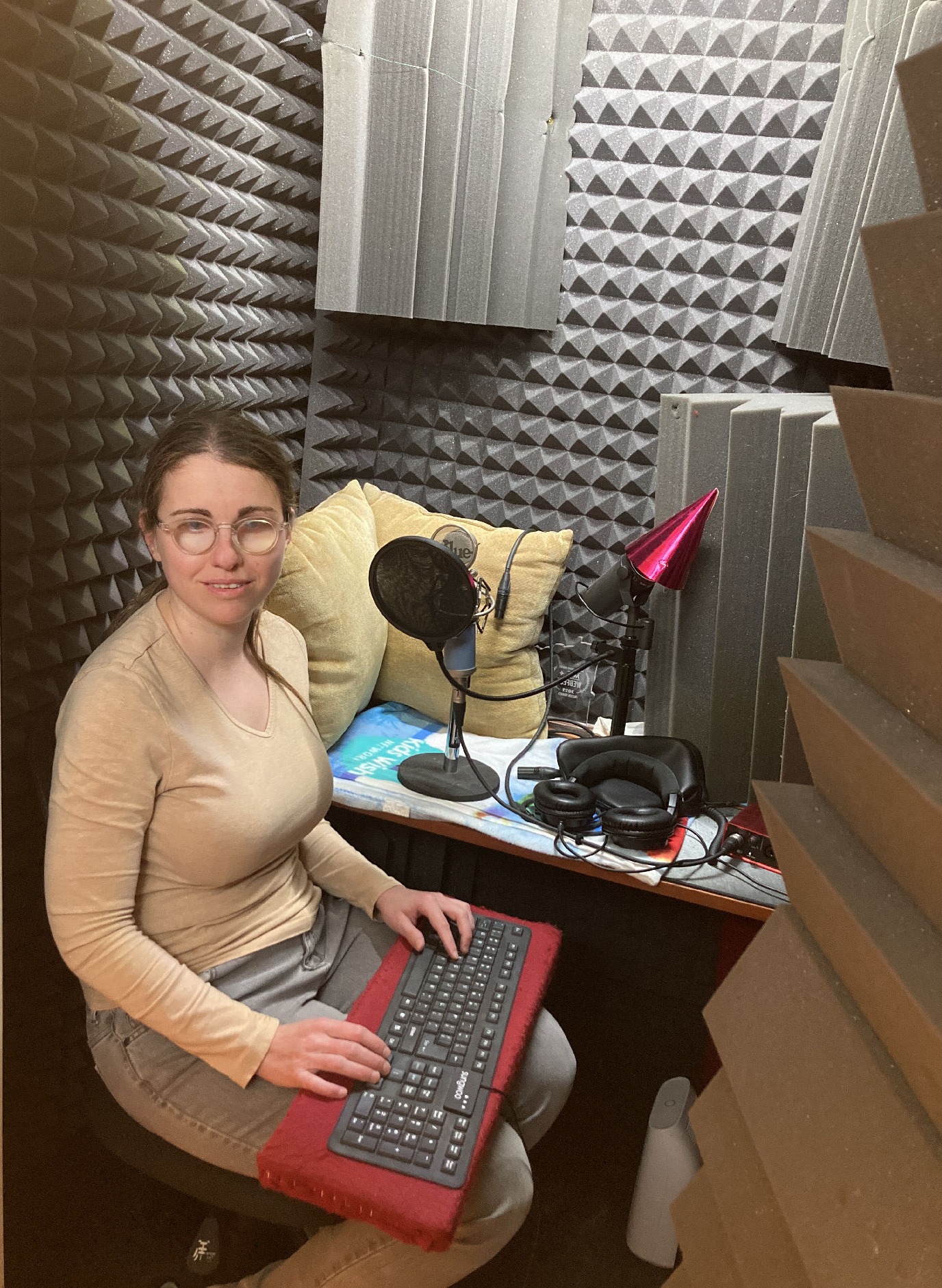 Tanja, co-founder of GetBraille - a Caucasian woman with brown hair tied in a ponytail - sits in a recording sound booth with grey soundproofing materials on its walls. She is wearing a tan long-sleeved shirt, grey jeans and rests her fingers on a silicon keyboard resting on a red cloth-covered wooden lap desk.