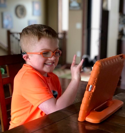 Dallas, a teen boy with CHARGE syndrome wearing a bright orange shirt and bright orange glasses, uses his iCanConnect-issued iPad, which also has a bright orange case