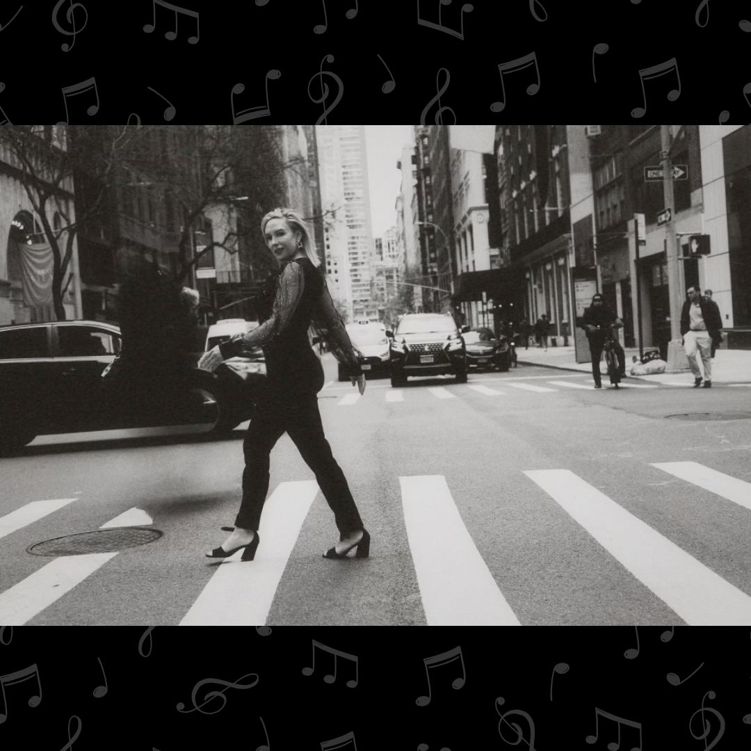 A black and white photo of recording artist Andrea Benham, a white woman with blonde hair, crossing an intersection in a busy city. She's dressed all in black, wearing black pumps and smiling at the camera.