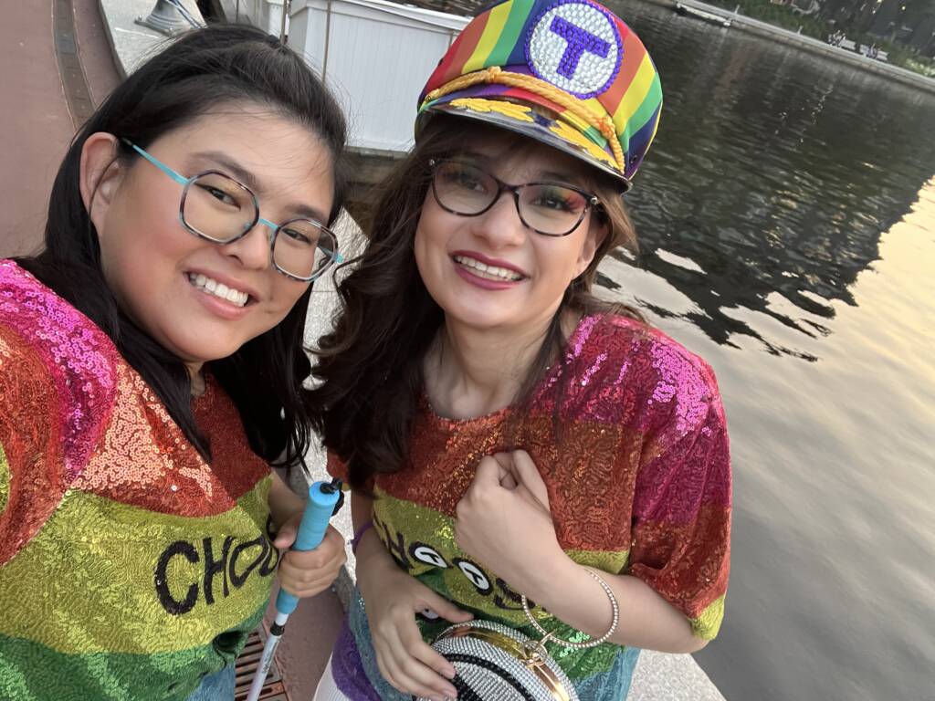 Tina and her girlfriend Pari, wearing matching sequined, rainbow-colored shirts that say, "Choose Love." smile for a selfie during a pride event.