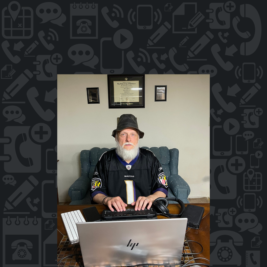Poet Matthew Harris sits at a desk, surrounded by technology - using a refreshable braille display, laptop computer and smartphone.