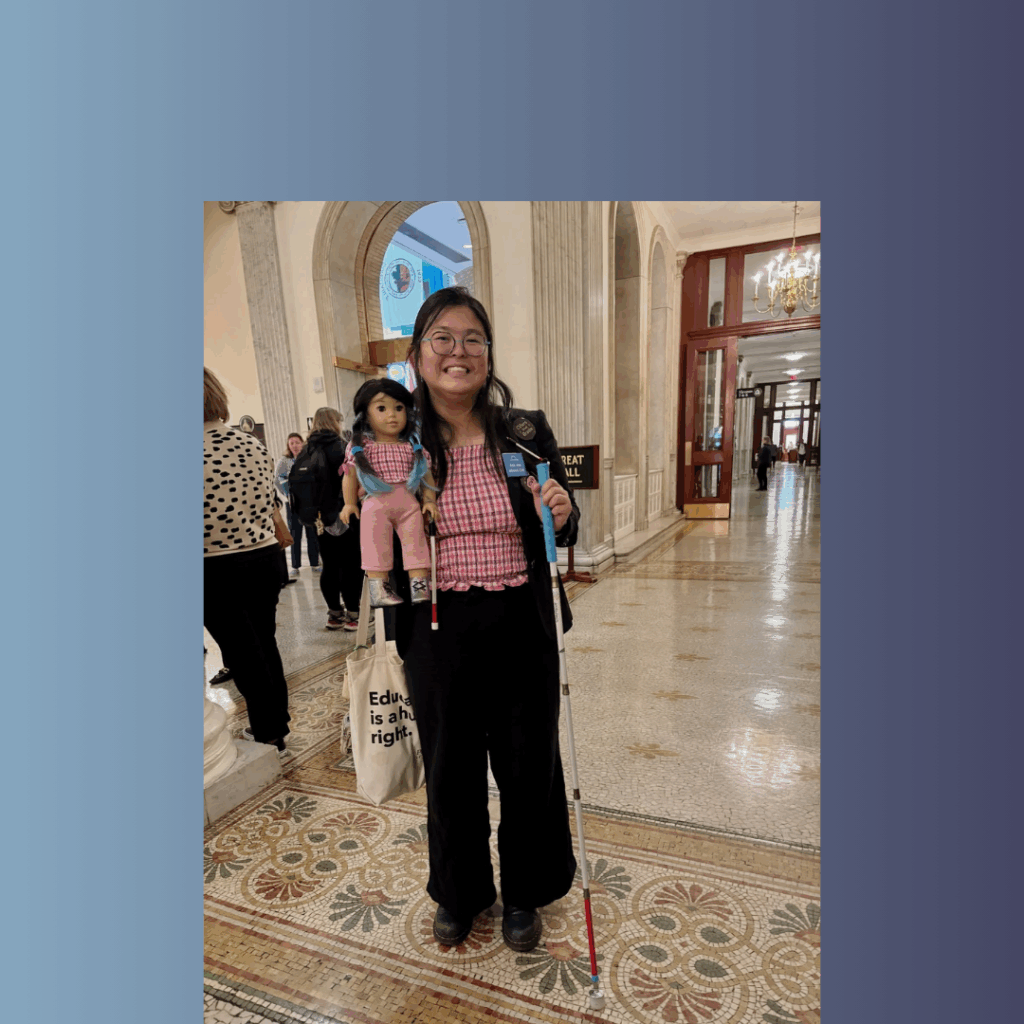Tina, a young Asian woman, smiles holding her white cane in one hand. In the other, she holds an American Girl doll that looks like an exact replica of her - right down to the matching outfit and doll-size cane.