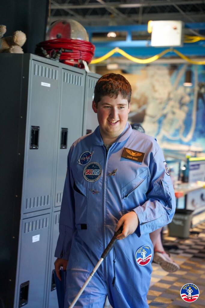 Declan, a teen boy, wearing a blue Space Camp space suit, uses his white cane to navigate through a classroom at camp