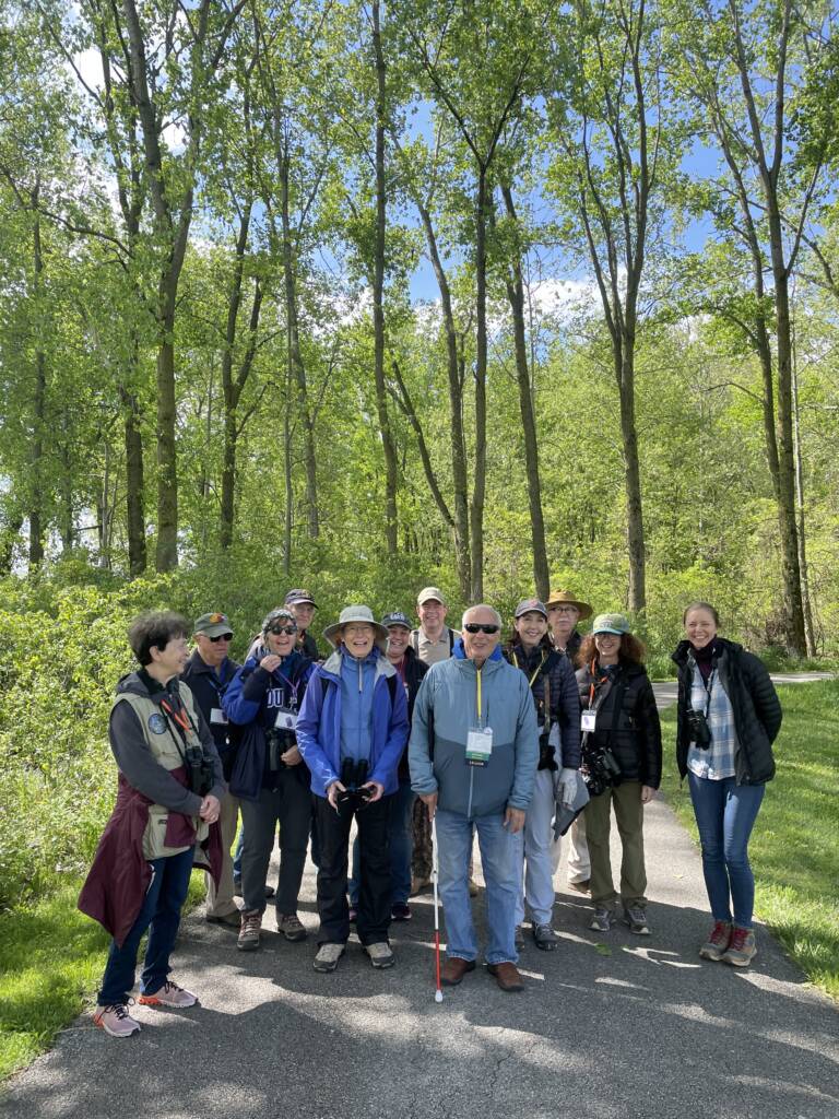 Jerry, holding his white cane, stands outdoors smiling at the front of a large group of fellow birders
