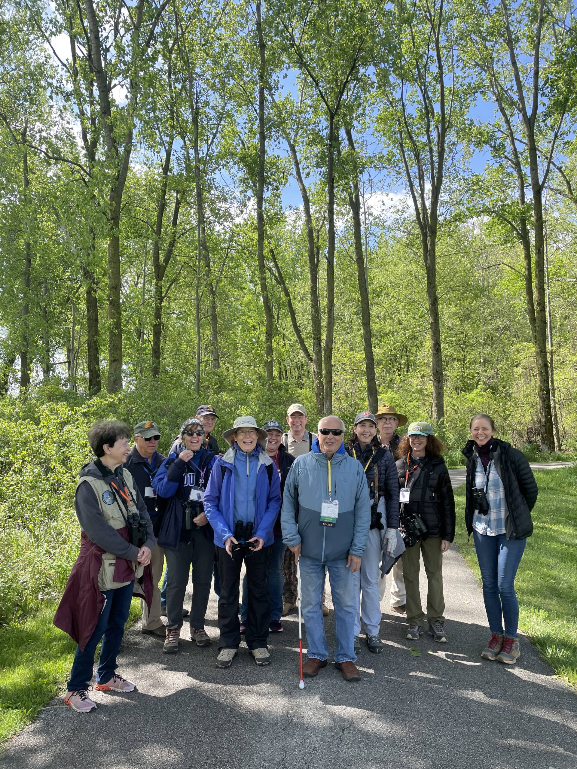 Jerry, holding his white cane, stands outdoors smiling at the front of a large group of fellow birders